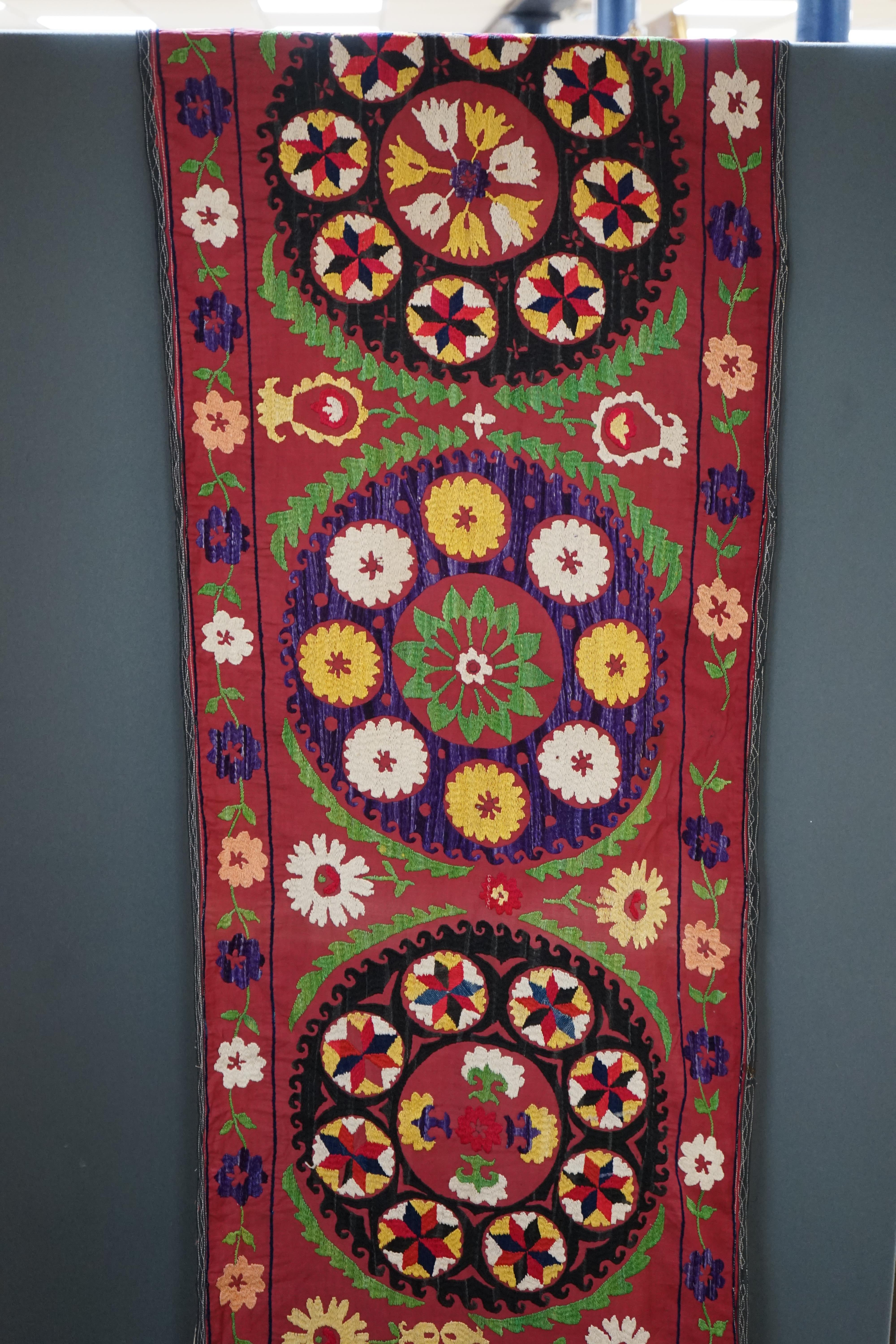 A 20th century Suzani wall hanging, possibly Uzbekistan, embroidered in bright polychrome silk threads into large symbols representing happiness, health and luck, on a burgundy cotton background, with a fine muslin backi